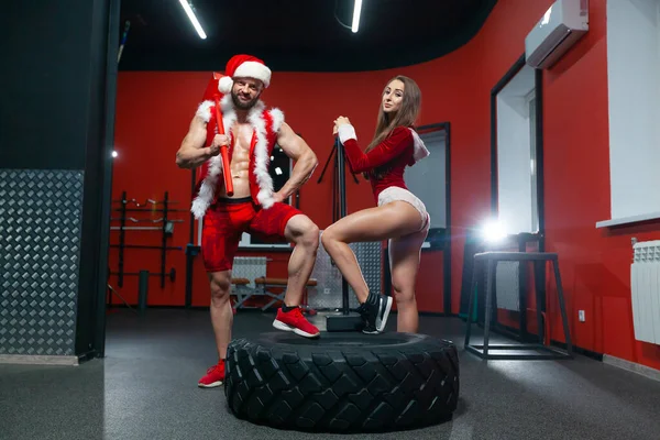 Christmas. Beautiful muscular man and woman in red Santa Claus costumes with hammers at the tire perform exercises in the gym.