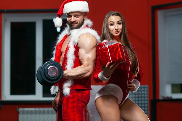 Karácsonykor. Jóképű, izmos férfi Mikulás jelmezben súlyzókkal és fitnesz nővel piros ruhában, ajándékkal. Edzőterem. — Stock Fotó