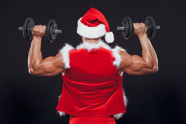 Sexy Santa Claus. Bodybuilder young handsome santa clause smile holds a dumbbells at New Years eve and Christmas winter holiday black background. The view from the back.