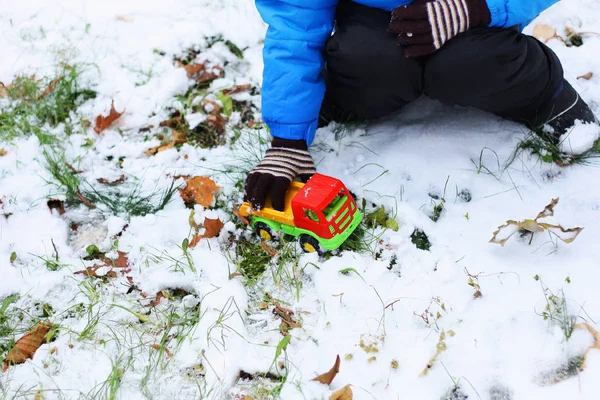 Children\'s winter games. The child sits among the first snow and plays with a red toy construction equipment. Visible hand, dressed in a blue jacket, black pants and boots.