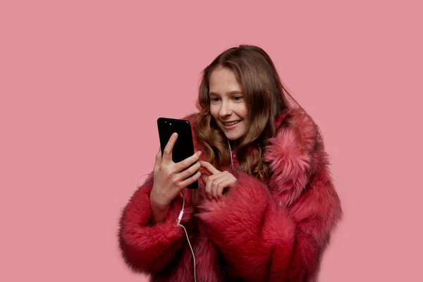 Mooi jong meisje schakelt liedjes op de telefoon in een roze modieuze bontjas — Stockfoto