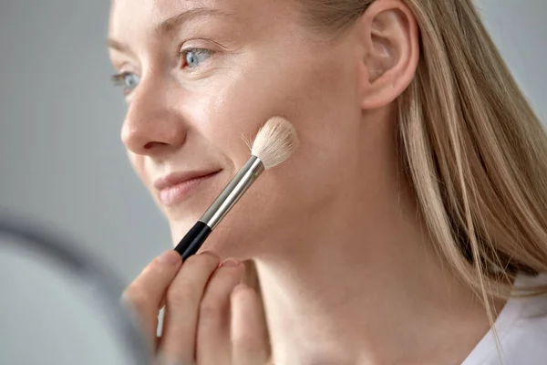 Um rosto grande de uma jovem que coloca um pincel cosmético em seu rosto . — Fotografia de Stock