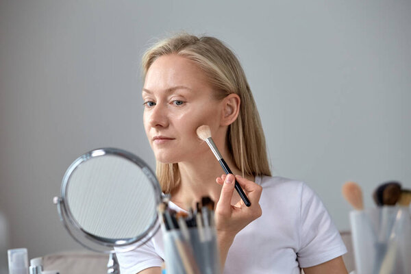 A beautiful blonde with bright blue eyes puts a makeup brush on her face.