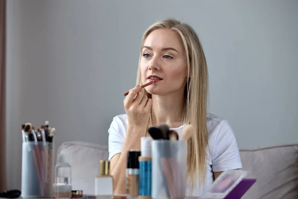 A blonde girl paints her lips with a pencil in front of the mirror of the house.