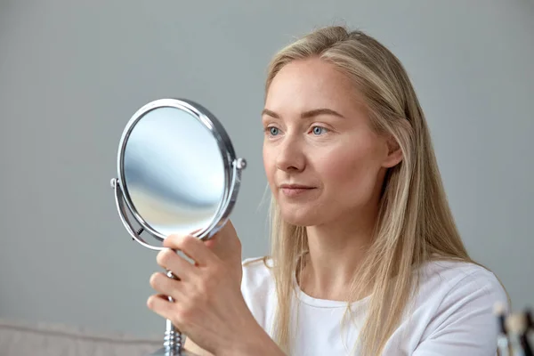 Woman natural blonde without makeup looks in a round mirror