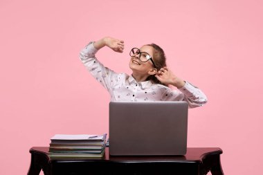 Genç bir kız büyük gözlüklerle gülümsüyor ve dizüstü bilgisayarın başında otururken yudumluyor..