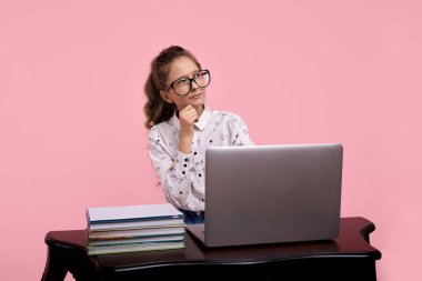 Masanın üzerinde bir dizüstü bilgisayar ve kitaplar var. Gözlüklü kız masada oturuyor..