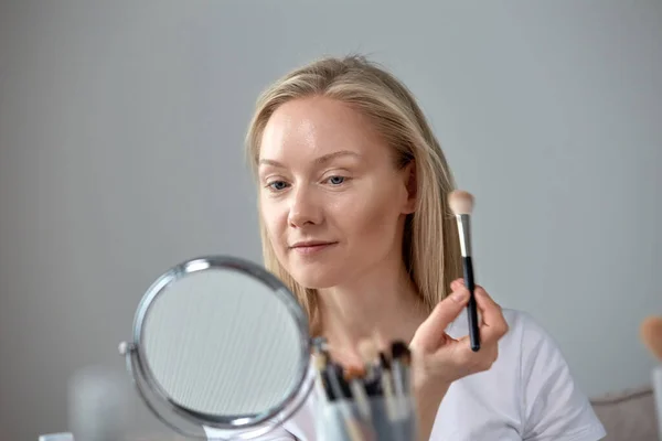 Una chica rubia con un cepillo de maquillaje en las manos. Una mujer se maquilla . —  Fotos de Stock