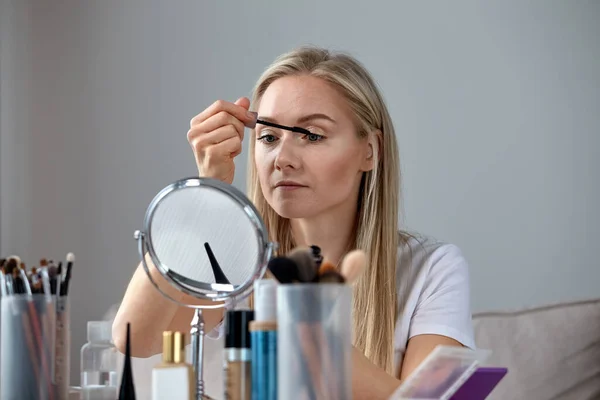 Een mooi blond meisje verft mascara. Grijze achtergrond. — Stockfoto