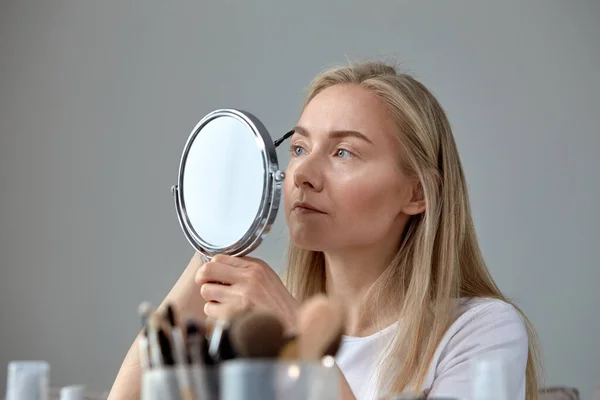 Молодая женщина смотрит в круглое зеркало и рисует брови карандашом . — стоковое фото