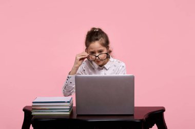 Masanın üzerinde bir dizüstü bilgisayar ve kitaplar var. Gözlüklü güzel bir kız oturuyor.