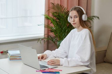 Kulaklık takan bir kız evde oturuyor. Masada dizüstü bilgisayarla oturuyor.