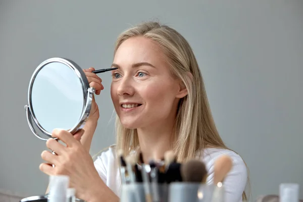 Een blonde vrouw maakt make-up voor de spiegel, tekent wenkbrauwen met een potlood — Stockfoto