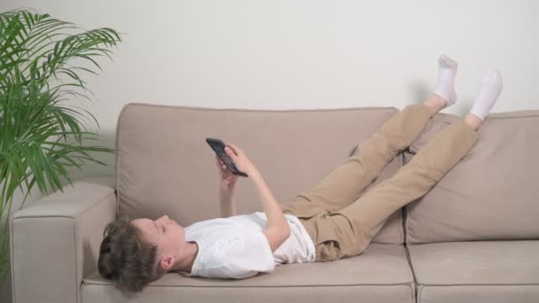 Der Junge liegt auf einem beigefarbenen Sofa, das Telefon in den Händen, die Beine hochgelegt. — Stockvideo