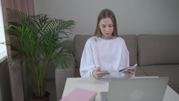 Ragazza seduta su un divano legge un libro in una copertina bianca. Sul tavolo c'è il portatile . — Video Stock