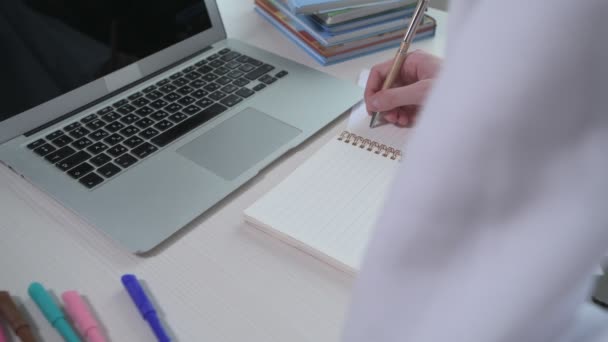 Das Mädchen schreibt in ein leeres Notizbuch. Auf dem Tisch liegt ein Laptop. Online-Kurse — Stockvideo