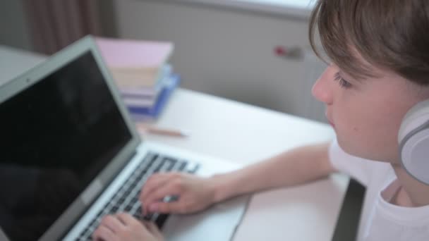 En pojke i hörlurar skriver på en laptop. markörer och böcker på bordet. — Stockvideo