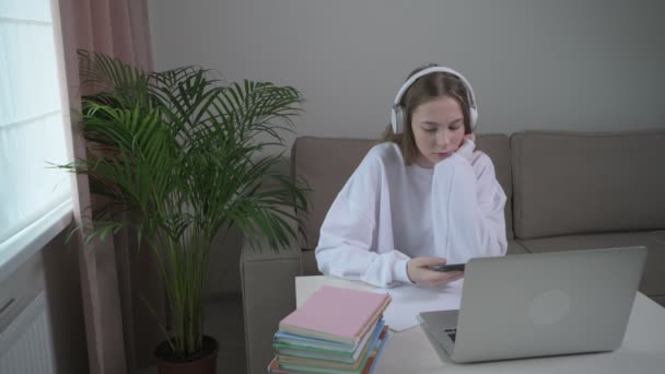 La fille écoute de la musique dans des écouteurs sans fil. Sur la table est un ordinateur portable — Video
