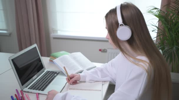 Flicka i trådlösa hörlurar skriver en föreläsning i en tom anteckningsbok. — Stockvideo