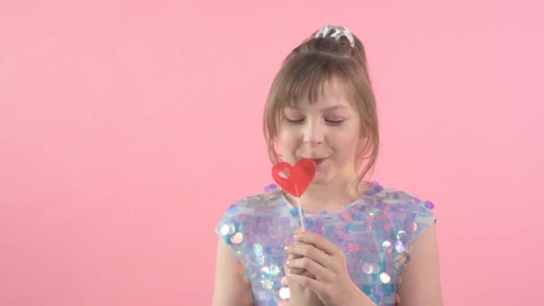 Gelukkig baby meisje in een helder jurk eten een hart-vormige zoet snoep op een stok. — Stockvideo