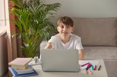 Masada oturmuş ödev yapan gülümseyen çocuk. Kameraya bak..