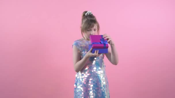 Egy csinos lány fényes ruhában kinyitja az ajándékot egy dobozban a születésnapjára.. — Stock videók