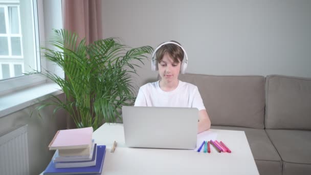 Egy fiú fejhallgatóban gépel egy laptopon. markerek és könyvek az asztalon. — Stock videók