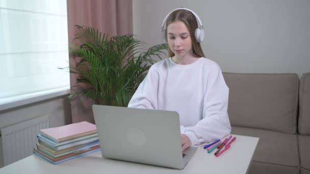 Ένα νεαρό κορίτσι ασχολείται με online μαθήματα με ακουστικά. Έπιπλα σπιτιού. — Αρχείο Βίντεο