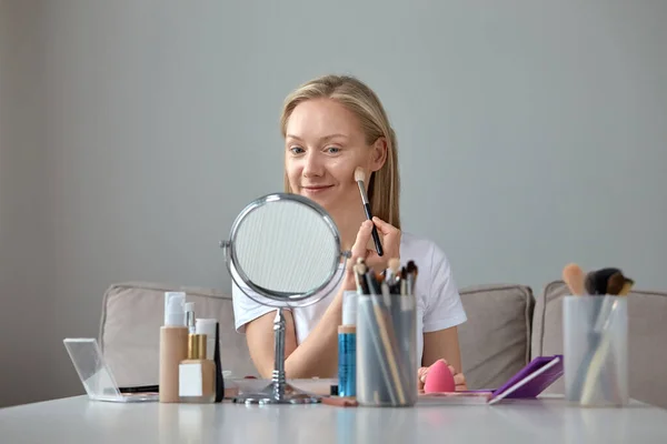 Μια νεαρή όμορφη ξανθιά κοπέλα κάνει make-up στο σπίτι μπροστά από έναν καθρέφτη. — Φωτογραφία Αρχείου