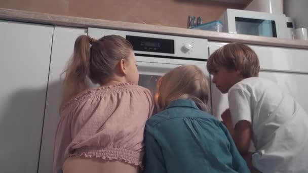 Niños rubios están sentados cerca del horno y esperando que los pasteles para cocinar . — Vídeos de Stock