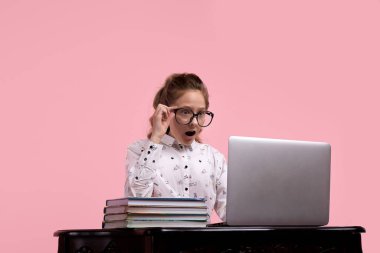 Şaşırmış bir kız elinde dizüstü bilgisayarla masada oturuyor ve tek eliyle bardakları tutuyor.