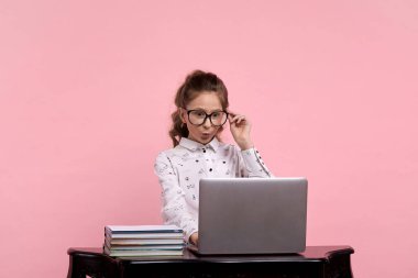 Pembe arka planda oturan kız şaşırmış yüzlü bir bilgisayarla masada oturuyor.