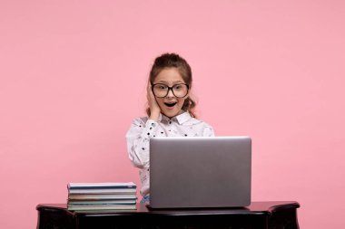 Şaşırmış bir kız elinde dizüstü bilgisayarla masada oturuyor ve tek eliyle bardakları tutuyor.