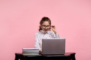 Masanın üzerinde bir dizüstü bilgisayar ve kitaplar var. Gözlüklü kız masada oturuyor..