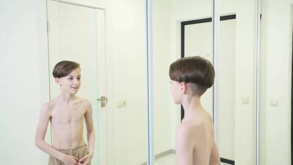 En tunn pojke står framför en spegel och beundrar sin fysiska styrka.. — Stockfoto