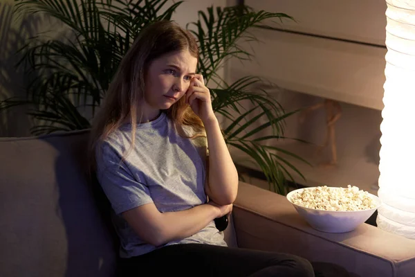 Una ragazza con la faccia annoiata sta guardando la TV. Popcorn nelle vicinanze. Camera accogliente . — Foto Stock