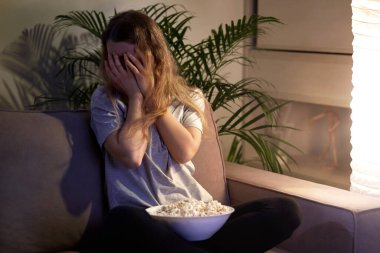 Korkunç yüzlü kız TV seyrediyor ve elleriyle yüzünü kapatıyor..
