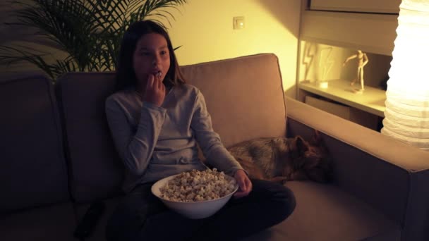 Bella ragazza dolce guarda la TV e mangia popcorn. Un simpatico cane dorme nelle vicinanze. — Video Stock