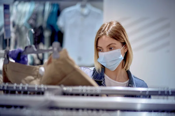 Ασφαλή ψώνια σε περίπτωση επιδημίας. Ένα κορίτσι με μάσκα εξετάζει ρούχα σε ένα κατάστημα.. — Φωτογραφία Αρχείου