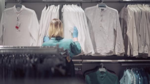 La ragazza bionda nel negozio sceglie i vestiti in una maschera medica e guanti. — Video Stock