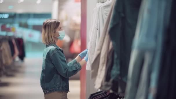 Vue latérale. Blonde fille dans un magasin de vêtements dans un masque médical et des gants. — Video