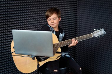 Bir çocuk gitarda şarkı söylüyor, gülümsüyor ve dizüstü bilgisayara bakıyor..