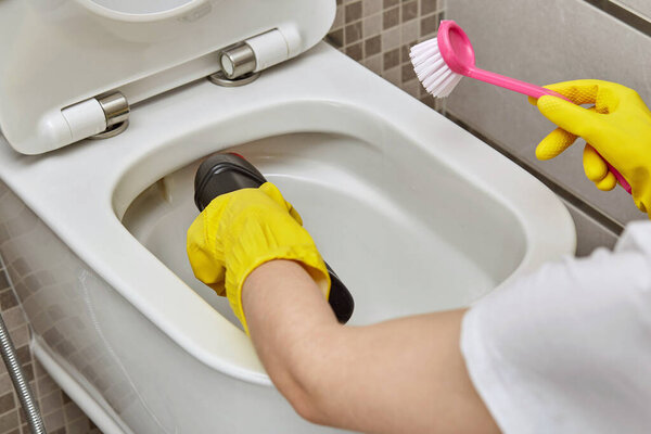 Hand in yellow rubber glove brushes the toilet bowl, pours household chemicals.