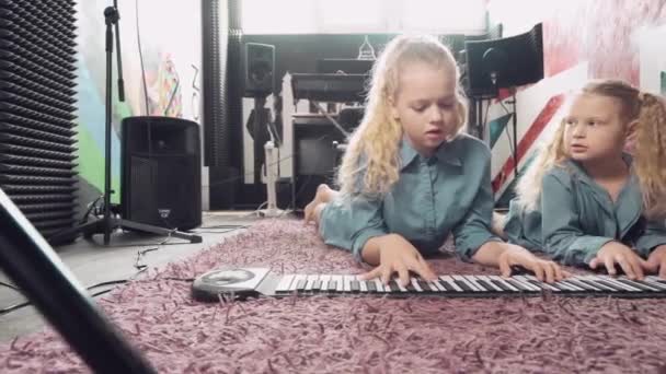 Twee leuke blonde meisjes spelen een muziekinstrument terwijl ze op een tapijt liggen. — Stockvideo