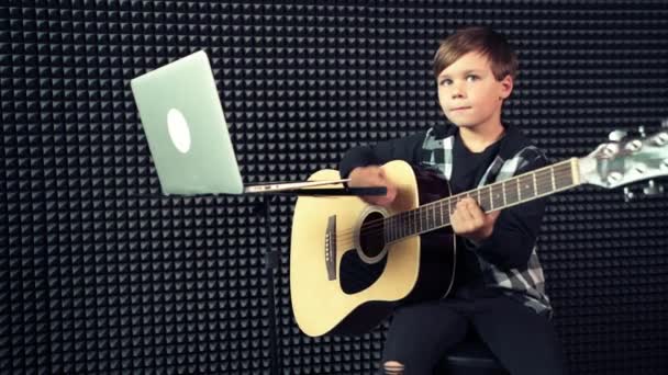 Primer plano de un niño tocando una guitarra acústica mientras está sentado en una silla . — Vídeo de stock