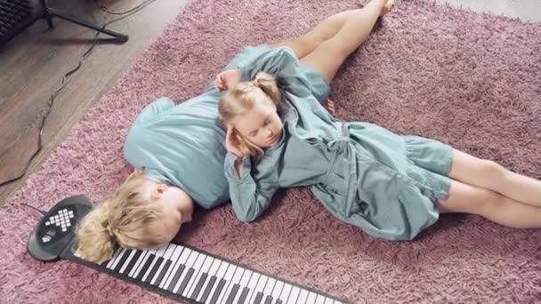 Vermoeide blonde meisjes ontspannen thuis op een roze tapijt. Vlakbij elektronische piano. — Stockvideo