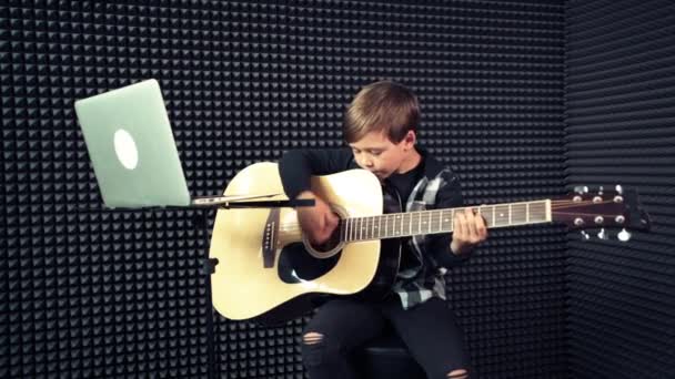 Primer plano de un niño tocando una guitarra acústica mientras está sentado en una silla . — Vídeo de stock