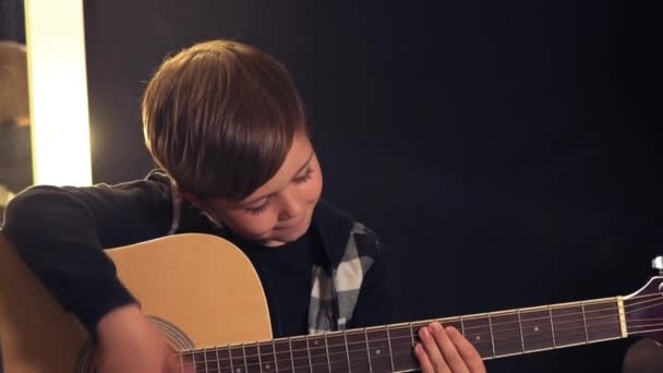 美しい笑顔のブルネットの少年はアコースティックギターを演奏しています。暗い背景. — ストック動画