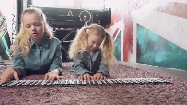Twee leuke blonde meisjes spelen op een synthesizer terwijl ze op de vloer liggen. — Stockvideo