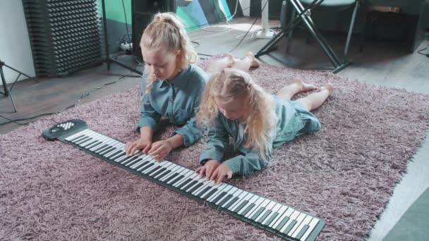Dwie blondynki grają na elektronicznym instrumencie, pianinie leżącym na różowym dywanie. — Wideo stockowe